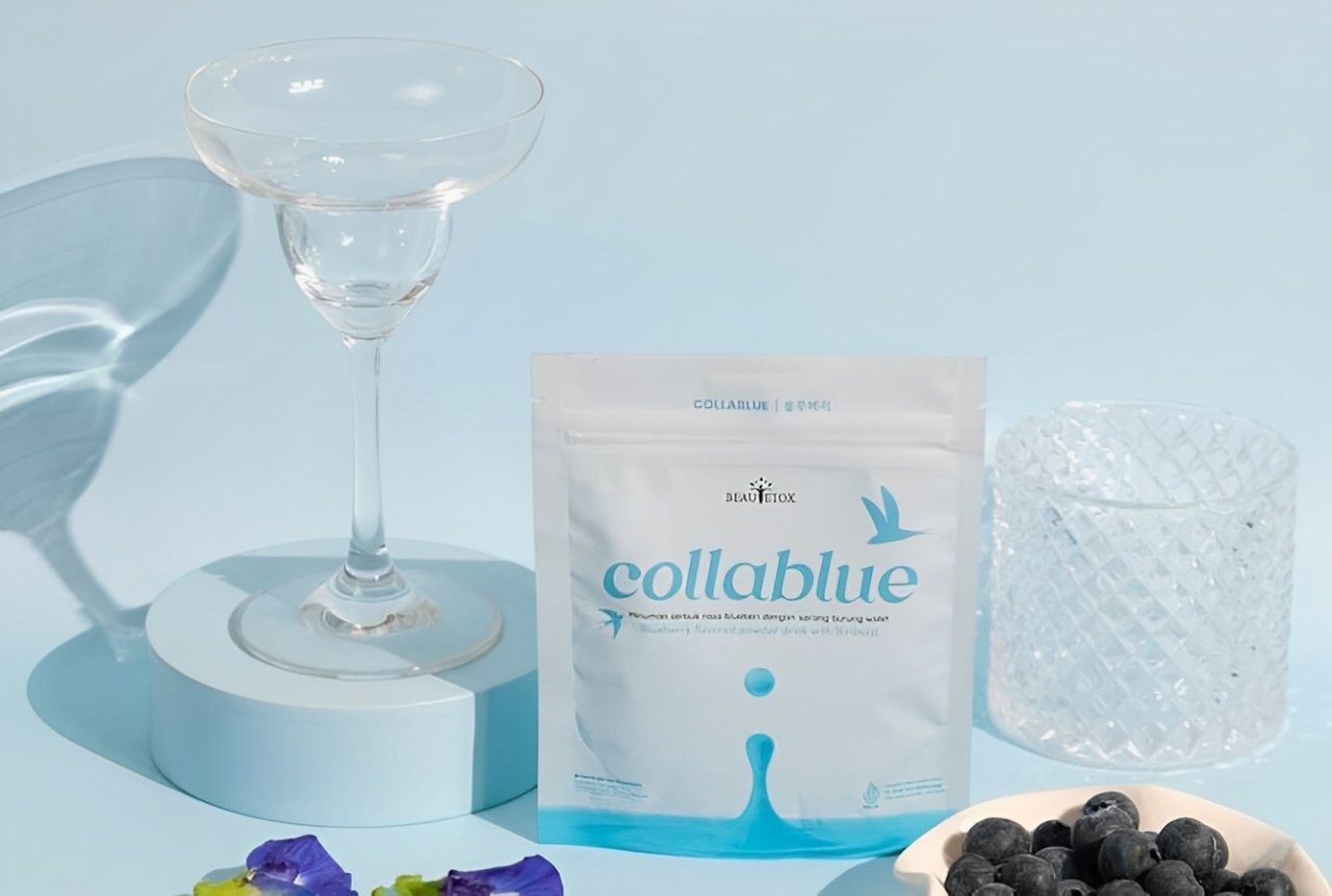 Produk minuman kolagen Beutox Collablue dalam kemasan putih dengan desain biru, dikelilingi oleh gelas kaca, blueberry, dan bunga telang.